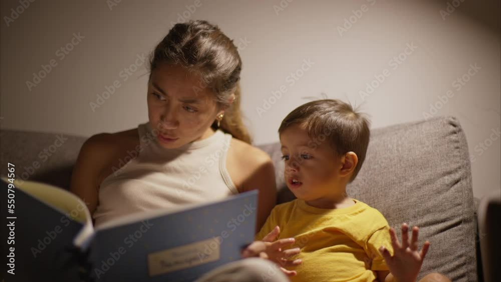 Cute brunette latin baby boy licking his fingers with chocolate cake while his mother reads a book to him sitting on a grey couch at night Cute brunette latin baby boy licking his fingers with chocolate cake while his mother reads a book to him sitting on a grey couch at night