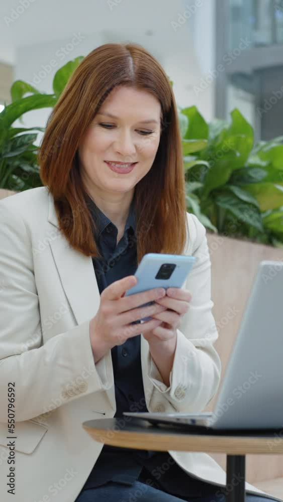 Vertical Screen: Cheerful businesswoman texting message on smartphone while sitting in cafe with laptop, online communication. Concept of technology Vertical Screen: Cheerful businesswoman texting message on smartphone while sitting in cafe with laptop, online communication. Concept of technology
