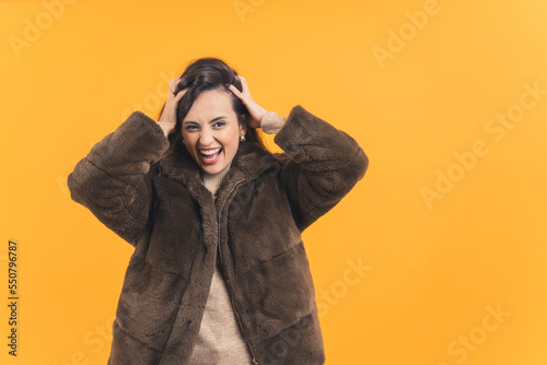 Young adult latin american woman wearing fur coat messing with her hair looking at camera with crazy laughing expression. Yellow background horizontal studio shot. High quality photo © PoppyPix