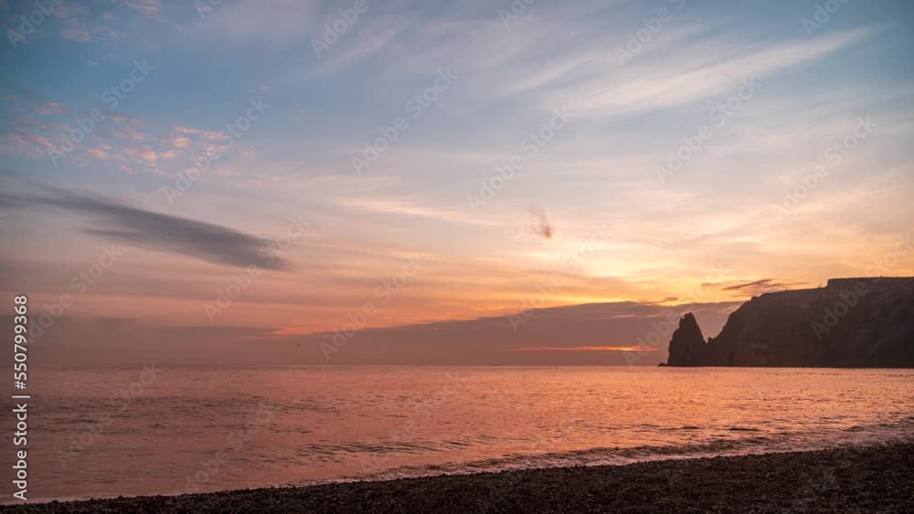 Sunset Sky Clouds Timelapse. A red burning sunset over the sea rocky volcanic cliff. Abstract nature summer ocean sea background. Small waves on golden warm water surface with bokeh lights from sun. Sunset Sky Clouds Timelapse. A red burning sunset over the sea rocky volcanic cliff. Abstract nature summer ocean sea background. Small waves on golden warm water surface with bokeh lights from sun.