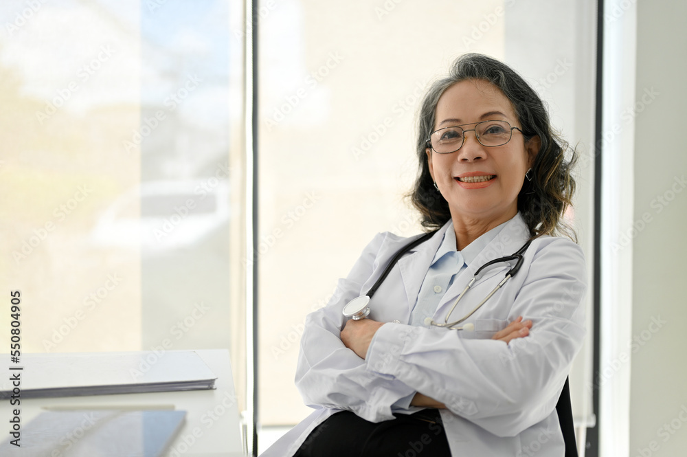 Successful Asian aged female doctor crossed arms and looking at camera while sitting by the window. Successful Asian aged female doctor crossed arms and looking at camera while sitting by the window.