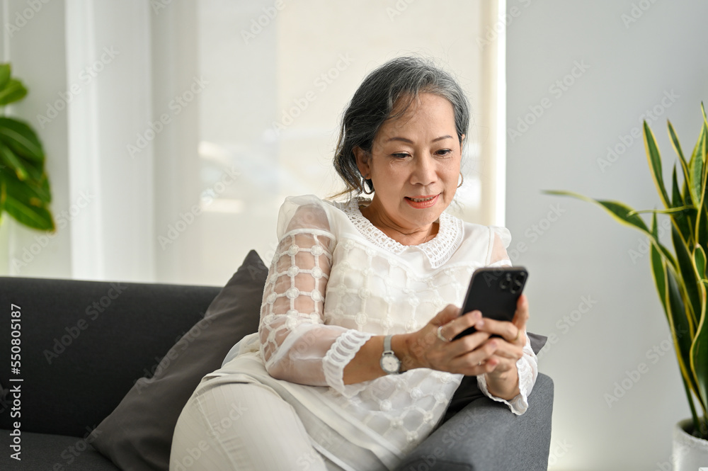 Happy Asian senior woman using her smartphone while relaxing on sofa in the living room. Happy Asian senior woman using her smartphone while relaxing on sofa in the living room.