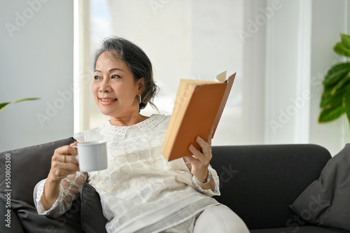 Happy Asian aged retire woman reading a book and sipping coffee while resting in her living room.