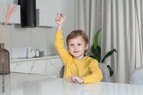Happy cute child boy in yellow sweater sitting by the table and raising his hand up with thumb up on white kitchen background