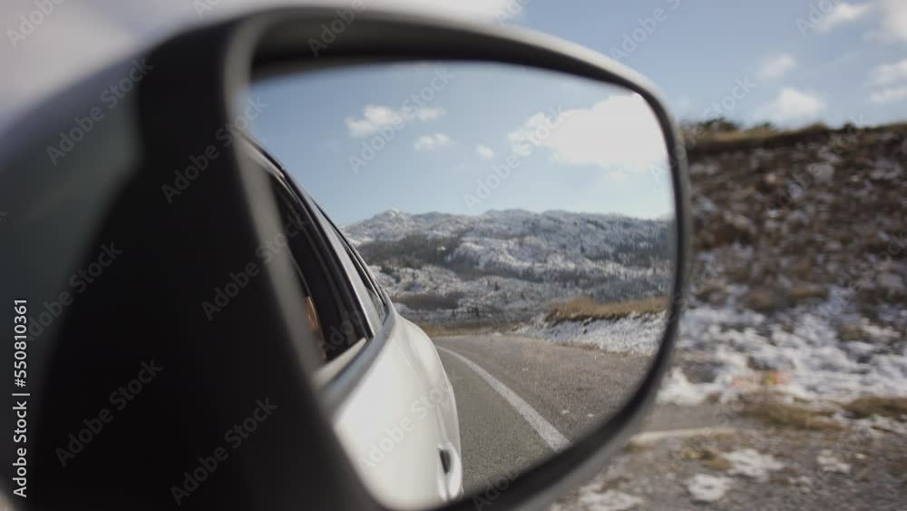 A passenger records a friend in the rearview mirror view of mountains in Alps and blue sky while driving with friends in the car A passenger records a friend in the rearview mirror view of mountains in Alps and blue sky while driving with friends in the car