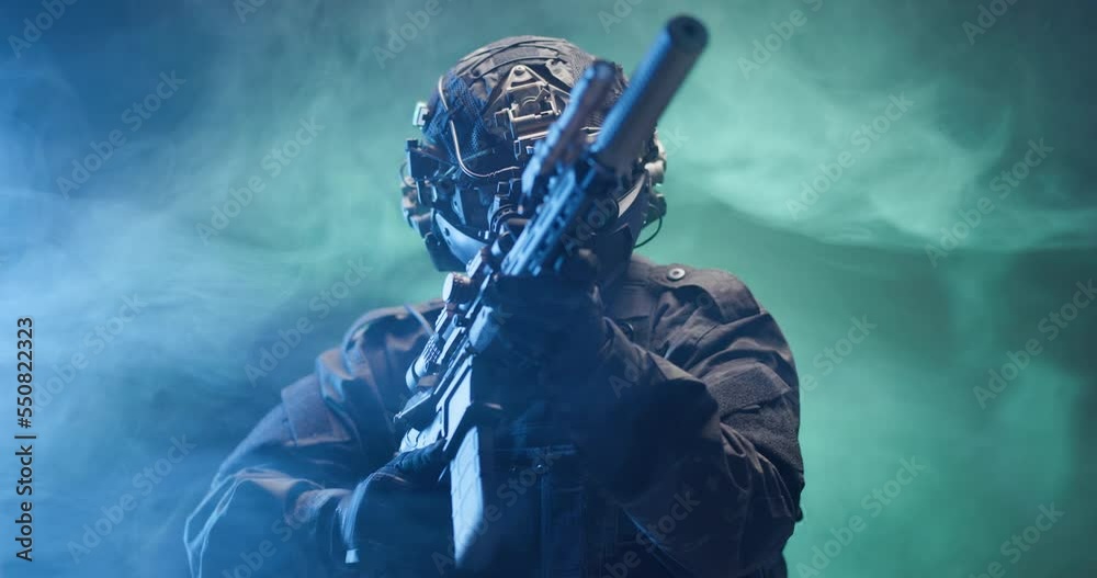 Soldier fully equipped with tactical gear and protection aiming his rifle at enemy with blue and green smoke in the background.