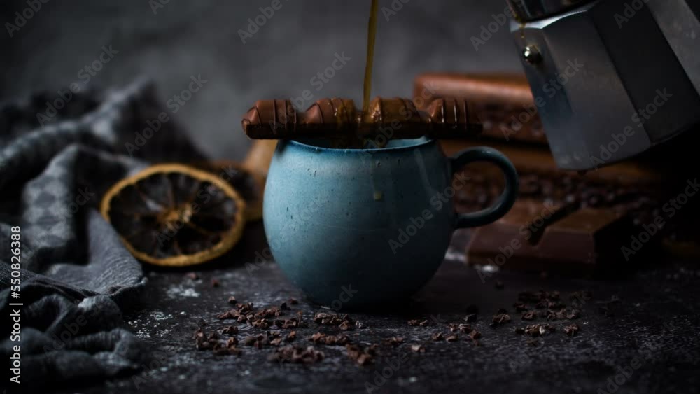 Pouring black coffee into blue vintage cup topped with melting chocolate bar from geyser coffee maker. Hot chocolate coffee in rustic barista scenery.  Chocolate and chocolate beans on dark table.