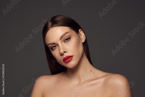 Portrait of female fashion model with naked shoulders and long hair looking at camera over dark grey background. Beautiful woman with trendy make-up, red lipstick and well-kept skin.