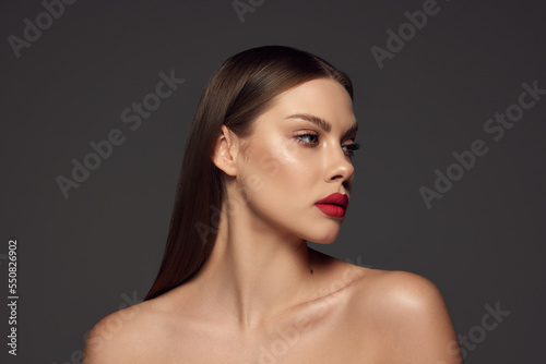 Portrait of female fashion model with naked shoulders and long hair looking away over dark grey background. Beautiful woman with trendy make-up, red lipstick and well-kept skin.