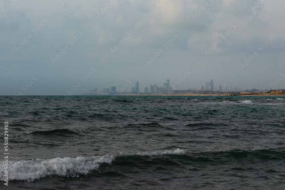Landscape photo of a cityscape beyond the sea shore. At the front is the blue ocean on a dark gloomy day