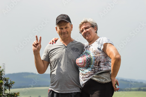 Happy senior couple tourists resting after hiking on top of the mountain. Gray sky background. Summer vacation journey.