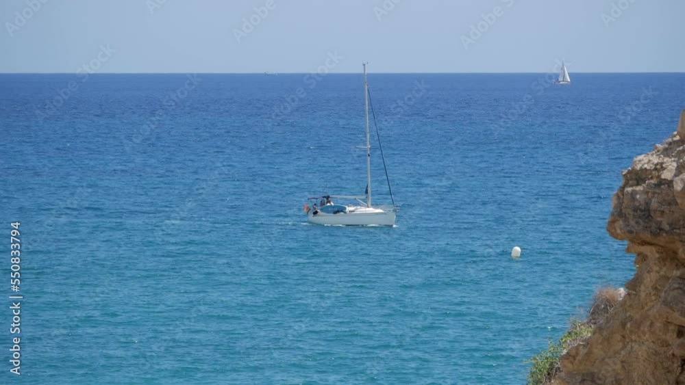 Close-up beautiful sailing boat on blue waters next to coast. Cliff rocks on view. Boat with mast but no sail navigates behind cliff. Other boat on distance. Holiday activity. Close-up beautiful sailing boat on blue waters next to coast. Cliff rocks on view. Boat with mast but no sail navigates behind cliff. Other boat on distance. Holiday activity.