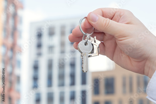 The man’s hand holds the key to the new apartment in the house in the background. The concept of renting a new apartment. Close up