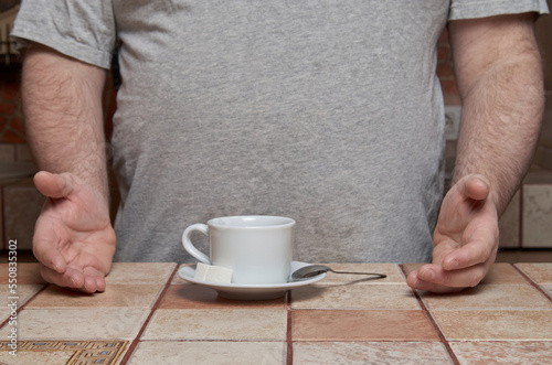 Human hands point to a coffee cup with a small spoon and sugar cubes on a saucer, standing on a tiled kitchen table