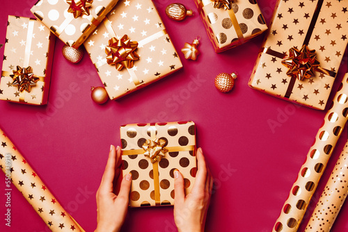Woman's hands holding gift box with golden starbow and ribbon on magenta background. Wrapping gifts concept. Christmas background. Top view. Color trend year 2023.