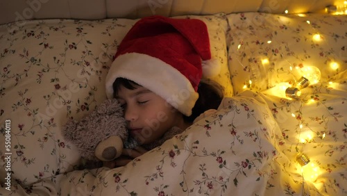 Little child sleep in bed with bear toy waiting for Santa Claus gifts before sleeping. Girl in red santa hat with lights on background New Years Eve, low light. 