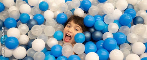 Banner happy little girl having fun in playroom. Kid lying on blue and white plastic balls in big dry paddling pool in playing centre. Portrait of child  smiling at camera. Leisure Activity.