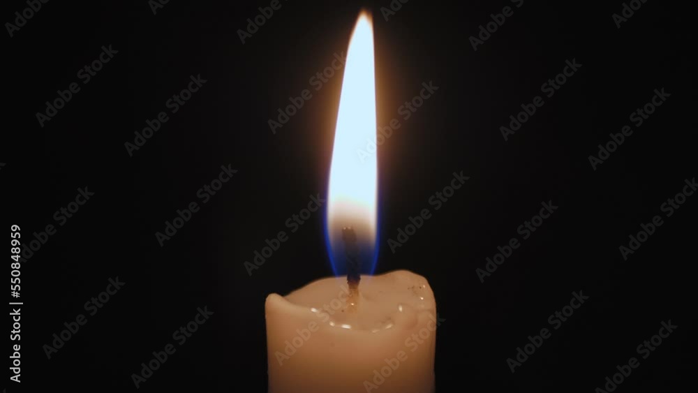 A burning candle close-up in a dark room, the flame from the candle illuminates the space around. Yellow-white fire from a candle. A burning candle close-up in a dark room, the flame from the candle illuminates the space around. Yellow-white fire from a candle.