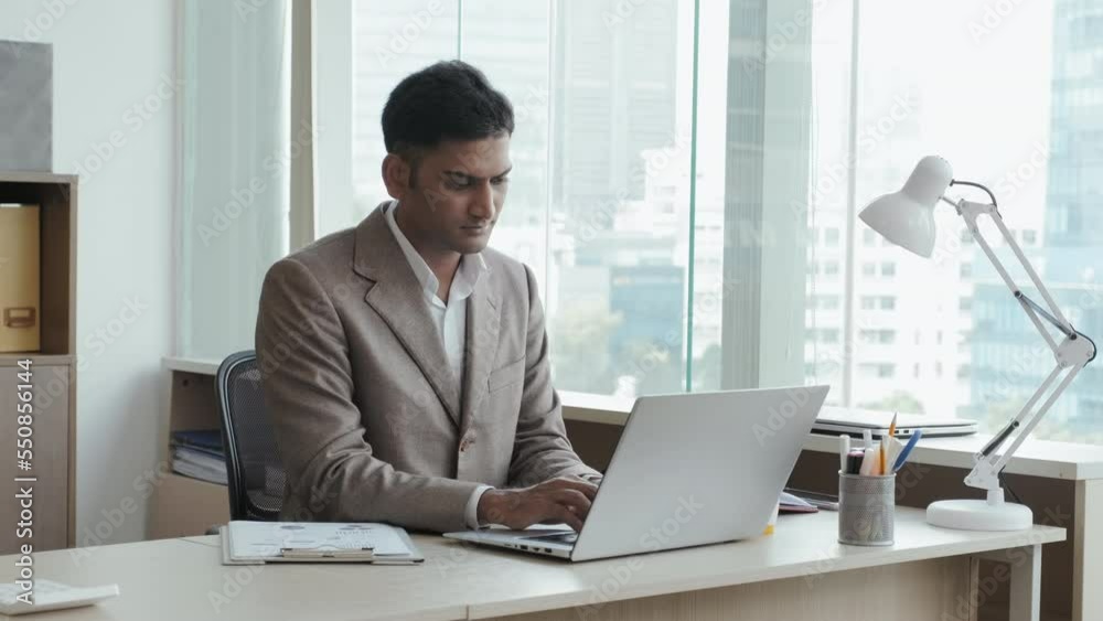 Medium shot of Indian businessman in formal suit working on laptop at office desk Medium shot of Indian businessman in formal suit working on laptop at office desk