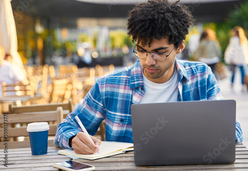 Pensive middle eastern student studying, taking notes, learning languages, online education concept. Young curly haired freelancer working online, planning project sitting at workplace 