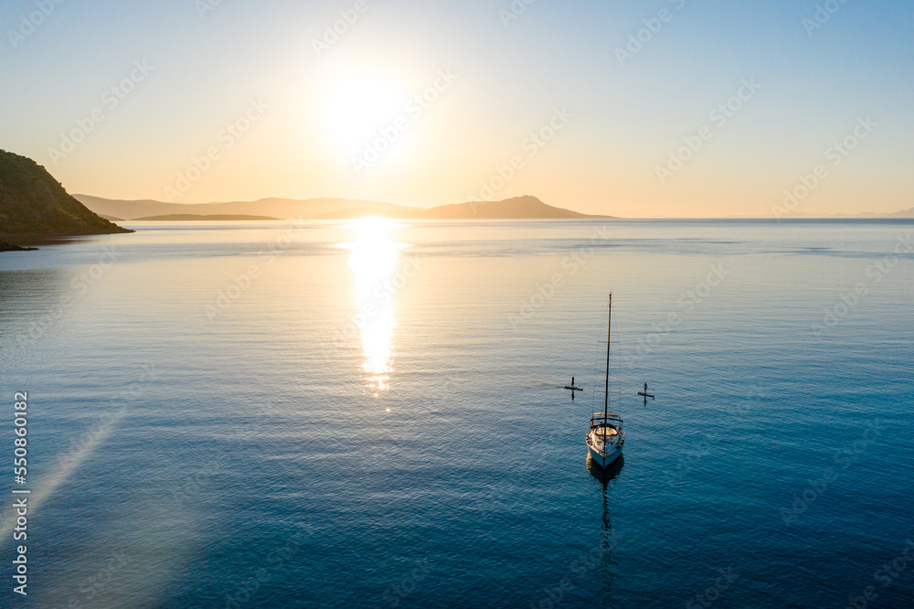 Aerial view of stand up paddlers women and a sailboat at sunset. Tranquil water of Mediterranean Sea. Aerial view of stand up paddlers women and a sailboat at sunset. Tranquil water of Mediterranean Sea.