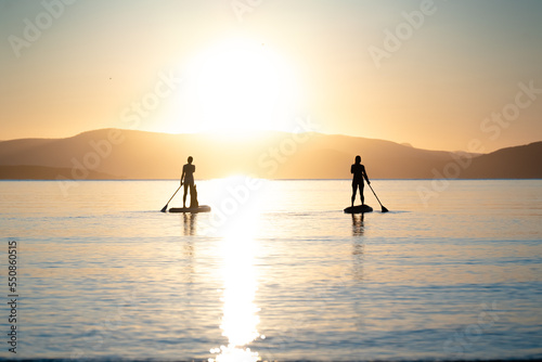 Silhouette of two stand up paddlers women at tranquil water of Mediterranean Sea at sunset.