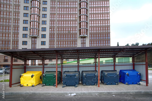 New colored plastic garbage bin for sorting various household waste. Colorful plastic bins for different waste under conopy next to new residential building. Place for dumpsters to collect garbage