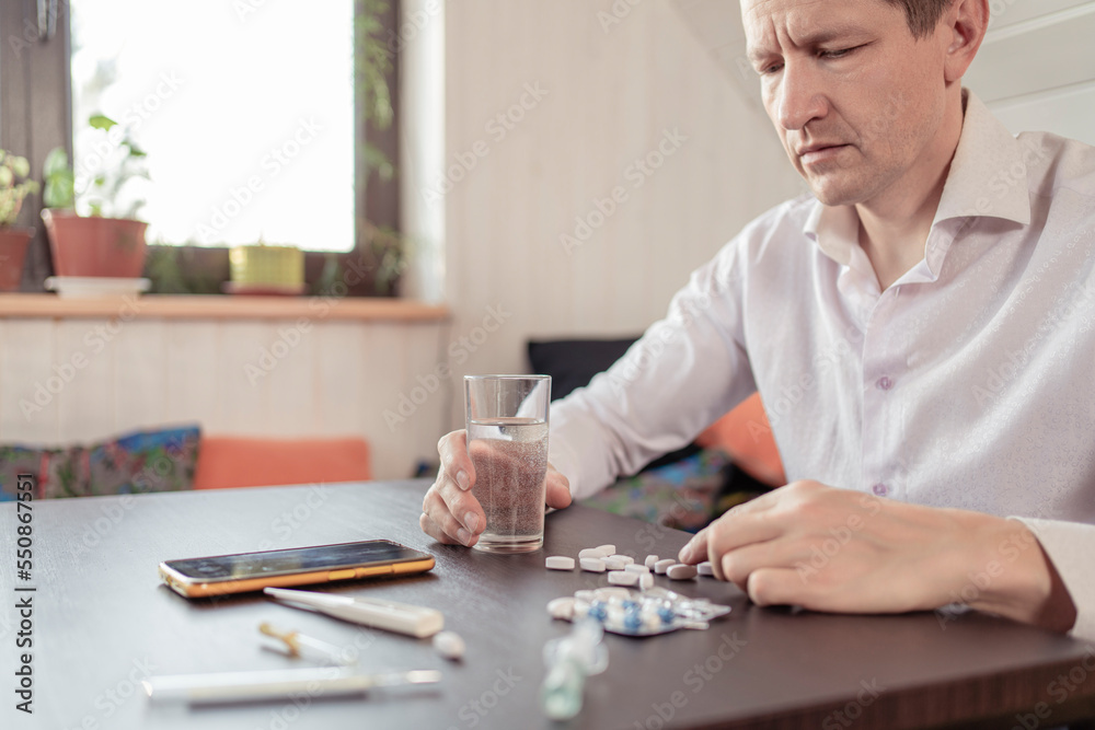 A sick person feels the symptoms of a viral disease, holds a thermometer in his hand. Tablets and water on the table.