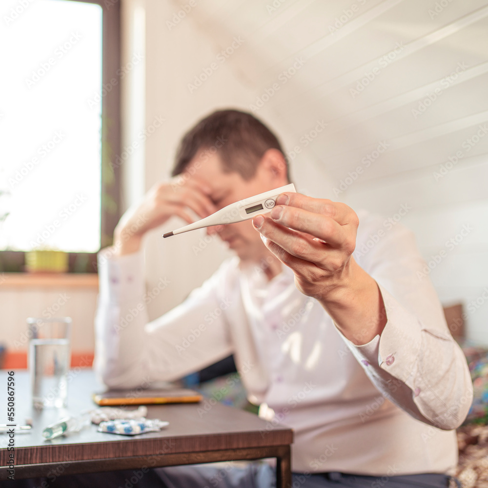 A sick person feels the symptoms of a viral disease, holds a thermometer in his hand. Tablets and water on the table. A sick person feels the symptoms of a viral disease, holds a thermometer in his hand. Tablets and water on the table.