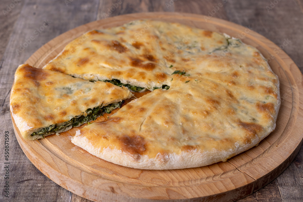 Pie with spinach, herbs and suluguni cheese on a wooden board. Whole pie with cut off piece. Traditional round flat pie with filling. Homemade of Caucasian and Balkarian cuisine