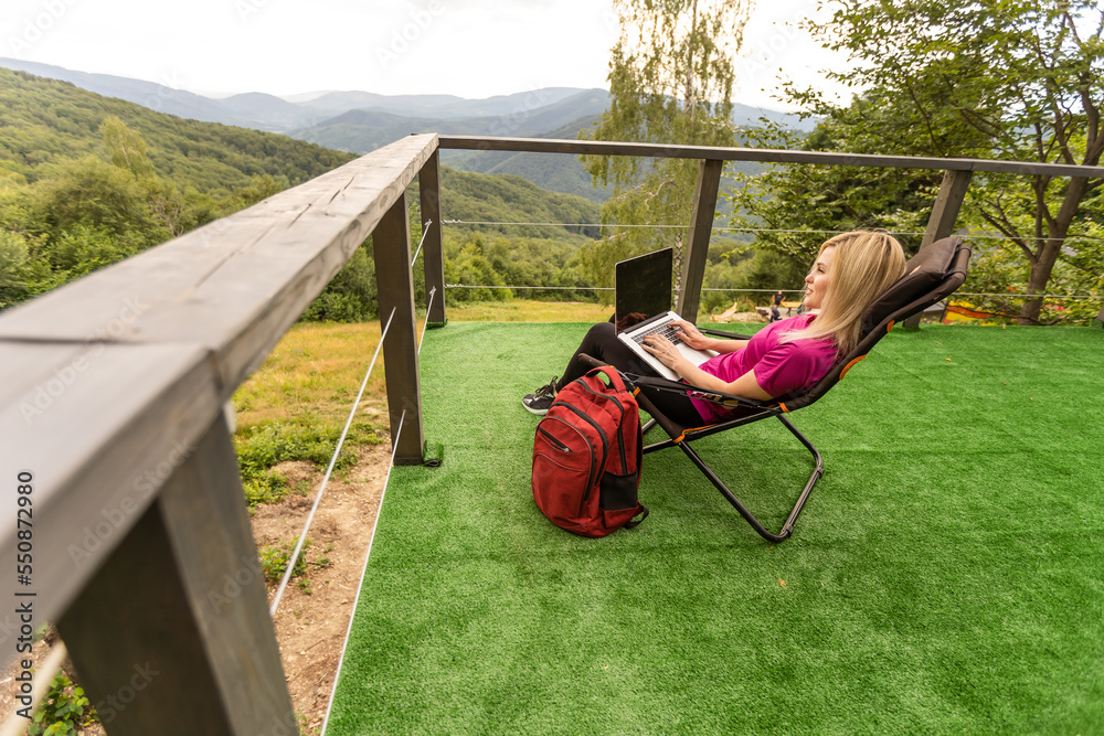 A woman in a sports warm suit works on a laptop outdoors in a mountainous area.
