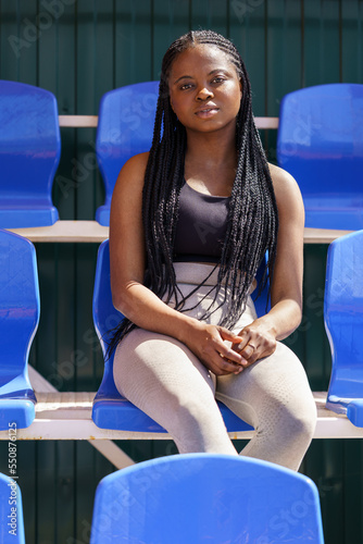 Ethnic African American girl high school student posing sits alone in seats for spectators at sports stadium. Youth black woman teenager in athletic clothes looking at camera with arms outstretched © DimaBerlin