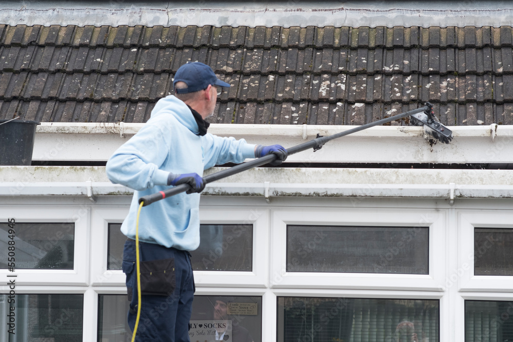 A window and gutter cleaner cleaning Dirty clogged white plastic pvc gutters and drain pipes with mossy green mould on plastic fascias. Blocked drains and guttering need regular yard work maintenance A window and gutter cleaner cleaning Dirty clogged white plastic pvc gutters and drain pipes with mossy green mould on plastic fascias. Blocked drains and guttering need regular yard work maintenance