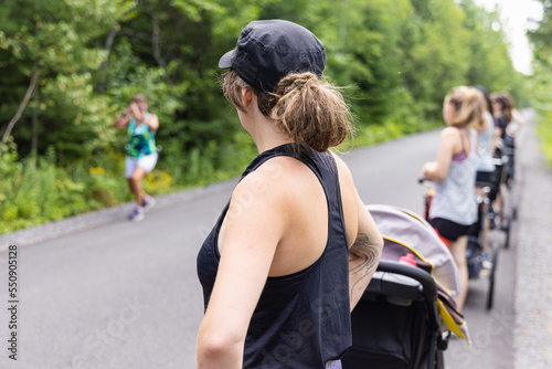 Shallow depth of field photo of woman standing on the side of a road, with other women and their strollers while trainer stands in front of the group photo