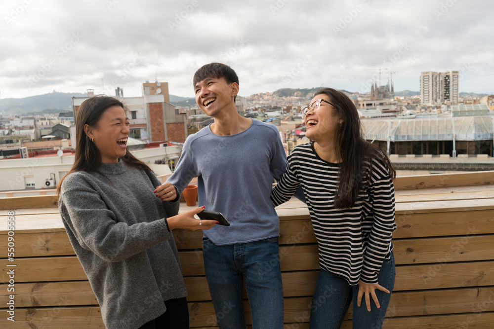 Three Chinese people standing on balcony and laughing loud while sharing smartphone.