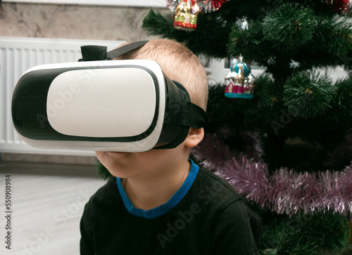 Obraz na płótnie A little boy in virtual reality glasses sits near a Christmas tree with toys. Close-up. w salonie