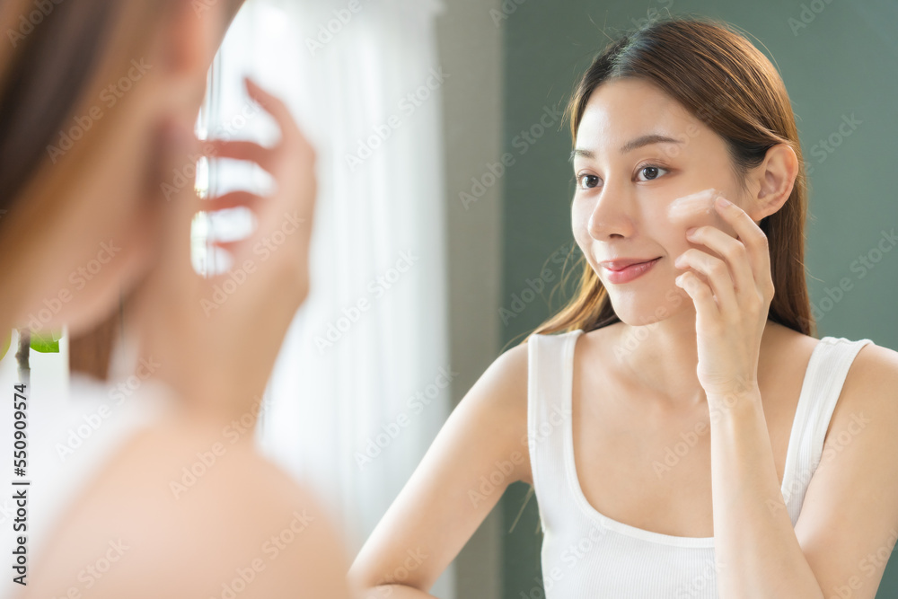 Fresh skin, beautiful smile of asian young woman, girl looking at mirror, hand applying moisturizer lotion on her face, putting cream treatment before makeup cosmetic routine at home. Facial Beauty. Fresh skin, beautiful smile of asian young woman, girl looking at mirror, hand applying moisturizer lotion on her face, putting cream treatment before makeup cosmetic routine at home. Facial Beauty.