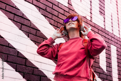 Stylish woman in magenta color jacket and heart shaped sunglasses wearing wireless headphones on her head and listening music. Urban city street fashion. Color of the 2023 year. Selective focus. photo