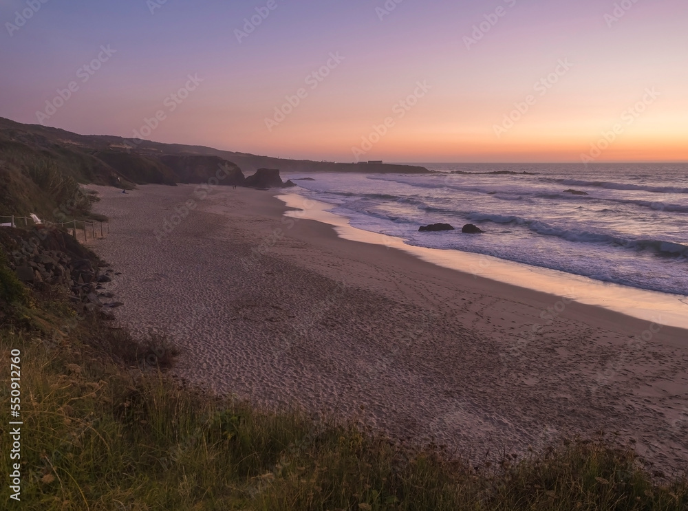 Sunset view of sand beach Praia Grande de Almograve with ocean waves in pink and red blue hour light, clear sky. Rota Vicentina coast, Almograve, Portugal