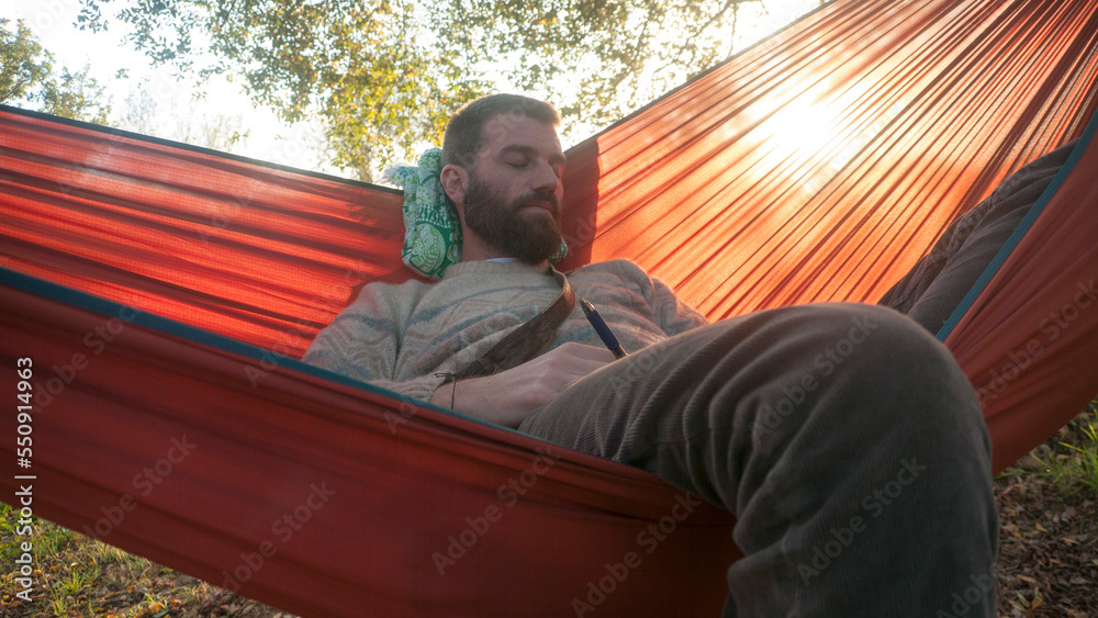 Fototapeta premium Hombre barbudo tumbado en hamaca entre arboles al atardecer