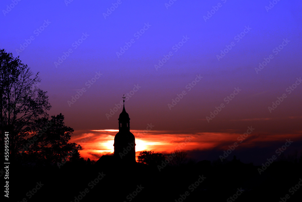 silouhette eines barocken kirchturms und eines labbaums vor ienem leuchtenden himmel bei sonnenuntergang