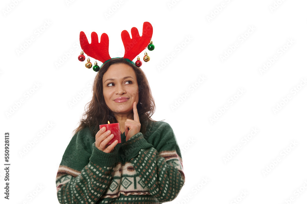 Isolated on white background, charming delightful multi-ethnic woman in warm Xmas patterned green sweater and deer antler hoop, holding a red lit Christmas candle and smiling looking up at copy space Isolated on white background, charming delightful multi-ethnic woman in warm Xmas patterned green sweater and deer antler hoop, holding a red lit Christmas candle and smiling looking up at copy space