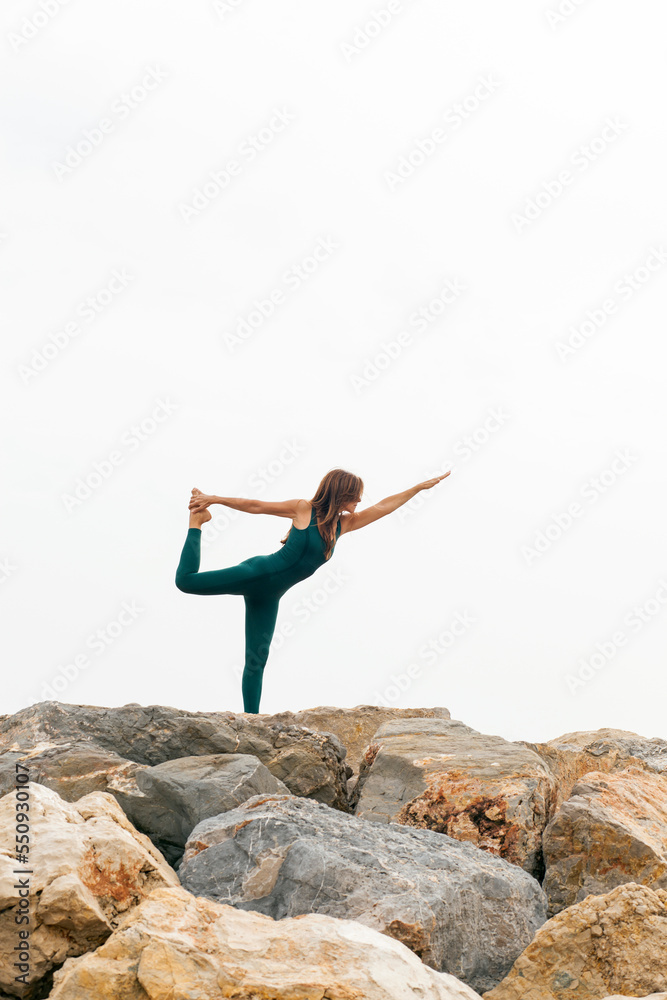 Beautiful woman standing in Natarajasana exercise, Lord of the Dance yoga pose (Lord Shiva’s) on a beach rock. Full length of sportive lady, working out. Relaxing and Mental health concept. Copy space Beautiful woman standing in Natarajasana exercise, Lord of the Dance yoga pose (Lord Shiva’s) on a beach rock. Full length of sportive lady, working out. Relaxing and Mental health concept. Copy space