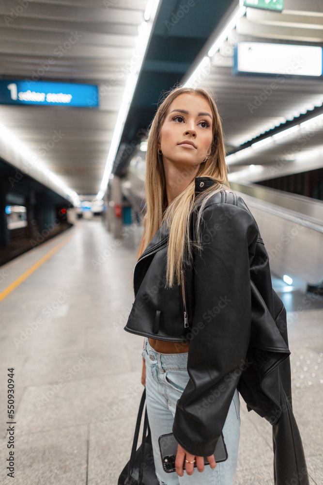 Stylish beautiful woman in a fashionable black rock leather jacket with blue jeans holds bag stands on a platform in the subway. Urban women's fashion and beauty