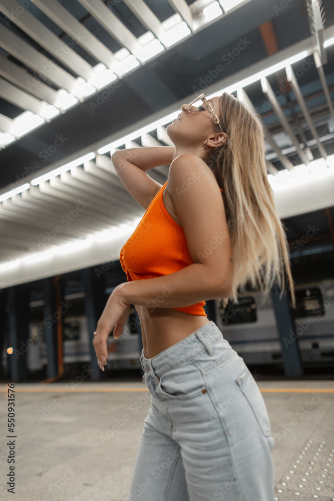 Fashionable beautiful urban hipster woman model with cool white sunglasses with body in fashion orange top with vintage jeans posing in subway Fashionable beautiful urban hipster woman model with cool white sunglasses with body in fashion orange top with vintage jeans posing in subway
