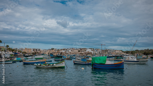 Luzzu, typical fishing boats of Malta moored in the marina of Marsaxlokk on an autumn day. Many ships in the bay, cityscape in the background.