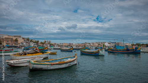 Luzzu, typical fishing boats of Malta moored in the marina of Marsaxlokk on an autumn day. Many ships in the bay, cityscape in the background.