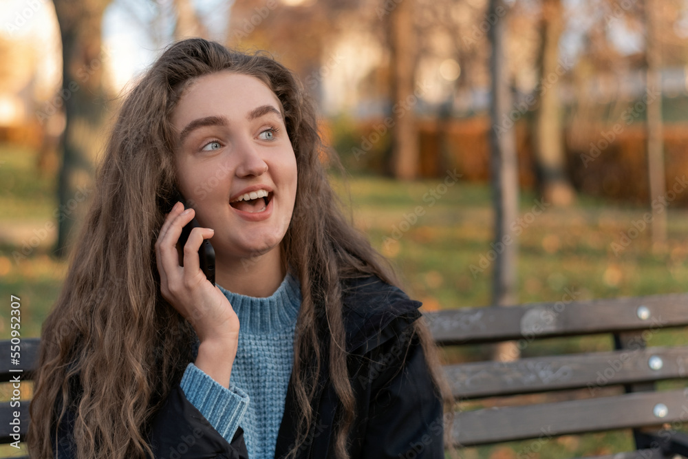 Cute teenage girl with long brown hair in park talking emotionally on phone. Young woman with smartphone outside. Cute teenage girl with long brown hair in park talking emotionally on phone. Young woman with smartphone outside.