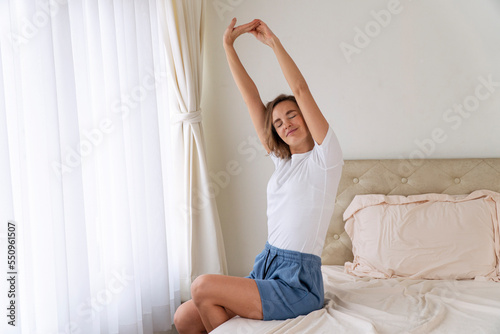 Activity caucasian slim woman doing stretch body raised arm after wake up sitting on bed in early beginner morning. Happy healthy female exercise stretching body laziness resting relax beside window.