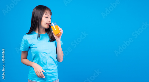 Enjoy asian woman drinking beverage freshness orange juice glass standing on blue background. Cheerful young female holding glass drink refreshment orange juice fruit for diet healthy with copy space.